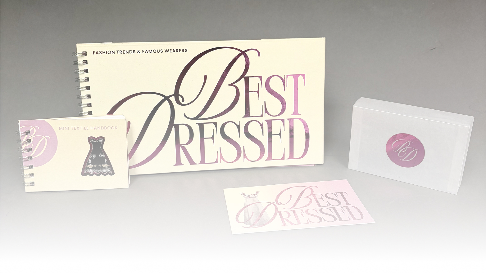 A long cream colored book with foiled pink letters that read fashion trends and famous wearers, BEST DRESSED, the book is spiral bound and sat next to a smaller textile handbook, a musuem donation card, and a vellum box for the mini textile handbook.